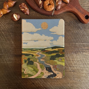 以下が含まれることがあります： 青い空と雲、金色の太陽の下、緑と黄色の野原を流れる川が描かれた風景をテーマにしたジャーナル。 ジャーナルは、焼き菓子が置かれた木製の表面にあります。