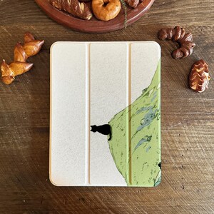 May include: A light beige tablet case with a black cat silhouette on a green and white illustrated design. The case is rectangular and folded. Baked goods are arranged on a wooden surface.