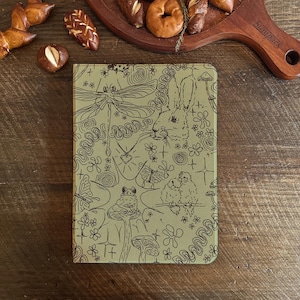 Puede incluir: Un diario verde con una ilustración caprichosa de una libélula, un conejo, mariposas y otros elementos inspirados en la naturaleza. La portada presenta un patrón repetitivo de flores y espirales.