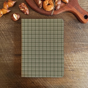 Pode incluir: Um caderno xadrez verde e bege está sobre uma superfície de madeira, rodeado por diversos produtos de panificação. A capa do caderno apresenta um padrão xadrez clássico, com pretzels, bagels e outros doces.