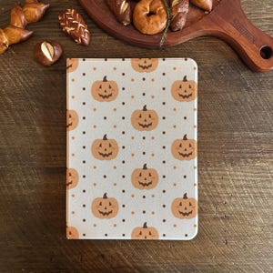 Pode incluir: Um pequeno caderno retangular com um padrão repetido de abóboras laranja e bolinhas marrons em um fundo cinza claro. O caderno está em uma superfície de madeira, com produtos de panificação ao fundo.