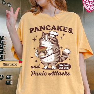 May include: A yellow t-shirt with a cartoon cat holding a stack of pancakes. The text on the shirt reads "Pancakes. and Panic Attacks. Buy One Get One".