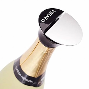 Può includere: Un tappo per bottiglia di champagne nero e argento con la scritta "AVINA" stampata sulla parte superiore. Il tappo è su una bottiglia di champagne con un'etichetta in foglia d'oro che dice "SANTA NEVADA".