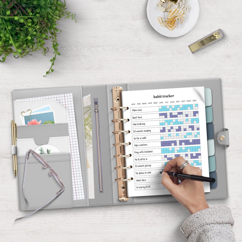 May include: A gray leather bound planner with gold rings and a habit tracker page. The habit tracker page has a list of habits to track, including "Make bed", "Quiet time", "20 minutes reading", "Go for a walk", "Wipe counters", "Pray with husband", "No to after 6", "10 minutes pickup", "No dishes in sink", "Wash face", "In bed by 10:30".