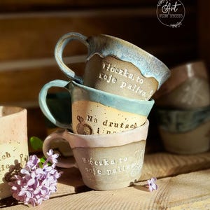 Może przedstawiać: Stos trzech ręcznie robionych ceramicznych kubków z tekstem w języku polskim. Kubki mają odcienie niebieskiego, beżowego i różowego i są ułożone na drewnianej półce. Każdy kubek ma uchwyt.