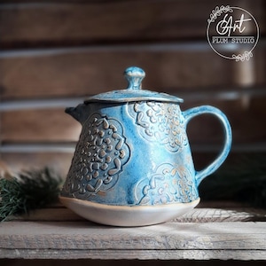 Könnte beinhalten: Eine blaue Teekanne aus Keramik mit weißem Sockel und goldenen Akzenten. Die Teekanne hat einen Deckel und einen Henkel und ist mit einem sich wiederholenden Muster aus stilisierten Blumen verziert.