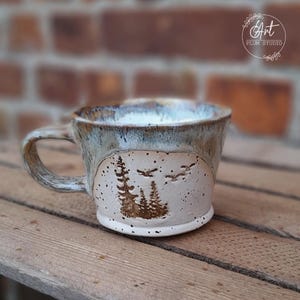 Könnte beinhalten: Eine weiße Keramiktasse mit brauner und blauer Glasur. Die Tasse hat eine Waldszene mit fliegenden Vögeln, die in die Seite geschnitzt sind. Die Tasse steht auf einer Holzoberfläche.
