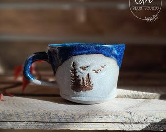 Może przedstawiać: Biały kubek ceramiczny z niebieskim rantem i miedzianym wzorem drzew i ptaków. Kubek stoi na drewnianej powierzchni.