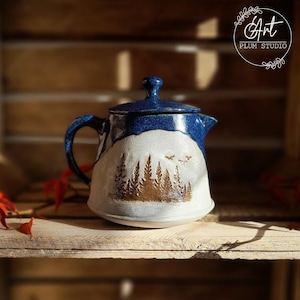Könnte beinhalten: Eine blaue und weiße Keramik-Teekanne mit einer Waldszene und Vögeln im Flug. Die Teekanne hat einen Deckel und einen Griff. Der Text "Art Plum Studio" ist auf dem Bild sichtbar.