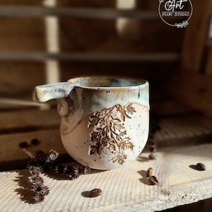 Può includere: Una tazza in ceramica fatta a mano con una glassa marrone chiaro, beige e blu. La tazza ha un'impugnatura unica e un disegno di felce sul lato. La tazza è appoggiata su una superficie di legno con chicchi di caffè sparsi intorno.