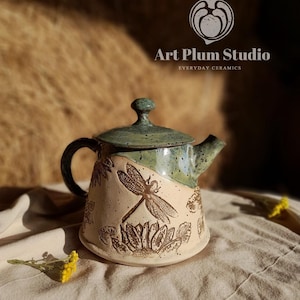 May include: A ceramic teapot with a speckled green lid and a beige base featuring embossed dragonfly and sunflower designs. The teapot has a dark handle and a spout. The text Art Plum Studio EVERYDAY CERAMICS is visible at the top.