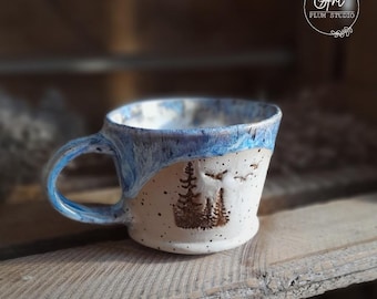 Może przedstawiać: Biały kubek ceramiczny z niebieską glazurą i brązową sylwetką drzew i ptaków. Kubek ma uchwyt i stoi na drewnianej powierzchni. Tekst "Art Plum Studio" jest widoczny w prawym górnym rogu obrazu.