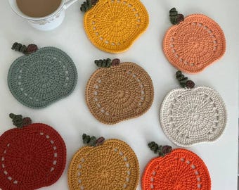 May include: A collection of handmade crochet pumpkin coasters in various fall colors, including orange, yellow, and brown. Each coaster features a stem and leaf detail, and a decorative openwork design. A cup of tea sits in the upper left corner.