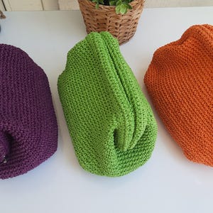 May include: Three handmade crochet clutch bags in purple, green, and orange. The bags are made of a textured yarn and have a rounded shape. The bags are displayed on a white surface.