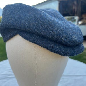 Puede incluir: Gorra de estilo newsboy de tweed azul. La gorra está hecha de una tela texturizada y tiene una corona redondeada y una visera corta y rígida. La gorra se muestra en una cabeza de maniquí.