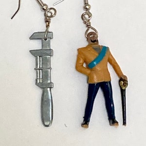 May include: A pair of earrings featuring a silver wrench and a miniature figurine of a man in a brown jacket, blue pants, and a blue sash. The man is holding a black cane.