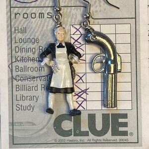 May include: A pair of earrings featuring a silver revolver and a miniature figurine of a maid in a black and white dress. The earrings are hanging from a card with the word "CLUE" and the text "©2002 Hasbro, Inc. All Rights Reserved. 00045".