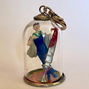 May include: A vintage keychain featuring a miniature glass dome. Inside, a figurine of a woman in a blue dress and a red lipstick are displayed. The dome has a silver base and a gold-colored clasp.