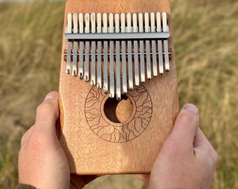 May include: A wooden kalimba, a plucked idiophone, held in hands. The instrument features metal tines of varying lengths and a sound hole with a sunburst design. The wood has a warm, natural tone, and the background is blurred.