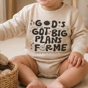 May include: A beige long-sleeve baby bodysuit featuring the black text "GOD'S GOT BIG PLANS FOR ME" with a small sun and smiley face graphic. The garment is worn by a smiling infant sitting near a woven basket containing a plush rabbit toy.