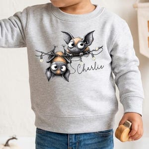 May include: A light gray long-sleeve sweatshirt featuring a graphic of two cartoon bats hanging from string lights. The name Charlie is printed in black script below the bats. The sweatshirt has a crew neckline and ribbed cuffs and hem.
