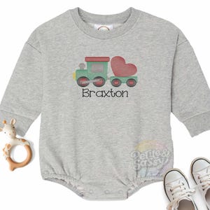 May include: A gray sweatshirt with a green and red embroidered train pulling a heart-shaped car. The name Braxton is embroidered below the train.