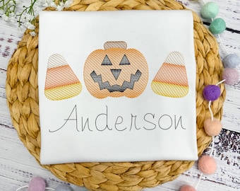 May include: White t-shirt with an embroidered Halloween design. The design features an orange jack-o'-lantern with a gray face, flanked by two candy corn pieces. The name "Anderson" is embroidered below the design.