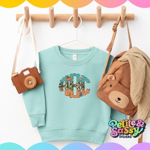 May include: A light blue sweatshirt with a fall-themed monogram design featuring a pumpkin, leaves, and a coffee cup. The monogram is surrounded by a leopard print pattern. The sweatshirt is hanging on a wooden rack with a brown backpack and a wooden toy camera.