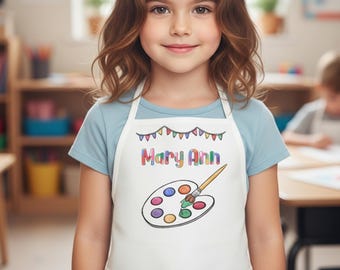 May include: A white apron with the name "Mary Ann" in colorful letters and a paint palette design. The apron is worn by a young person in a classroom setting. The apron has a bib and ties around the waist.