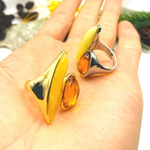 May include: Two rings featuring smooth, teardrop-shaped amber stones. One ring has a polished gold-tone band with a bright yellow amber piece and a smaller, translucent orange amber stone. The second ring has a silver-tone band with a pale yellow amber piece and a smaller, translucent orange amber stone.