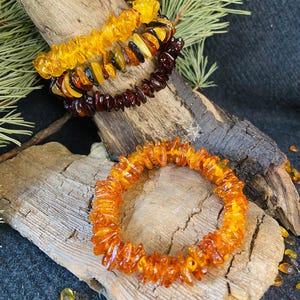 May include: Three amber bracelets with varying shades of yellow and brown are displayed on a piece of driftwood. The bracelets are made of irregular, polished amber stones strung together. A fourth bracelet, also amber, is in the foreground.