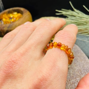 May include: A ring made of amber beads. The ring is a warm, golden-orange color and appears to be made of small, irregular-shaped beads. The ring is worn on a finger. The background is blurred, but includes a wooden bowl and greenery.