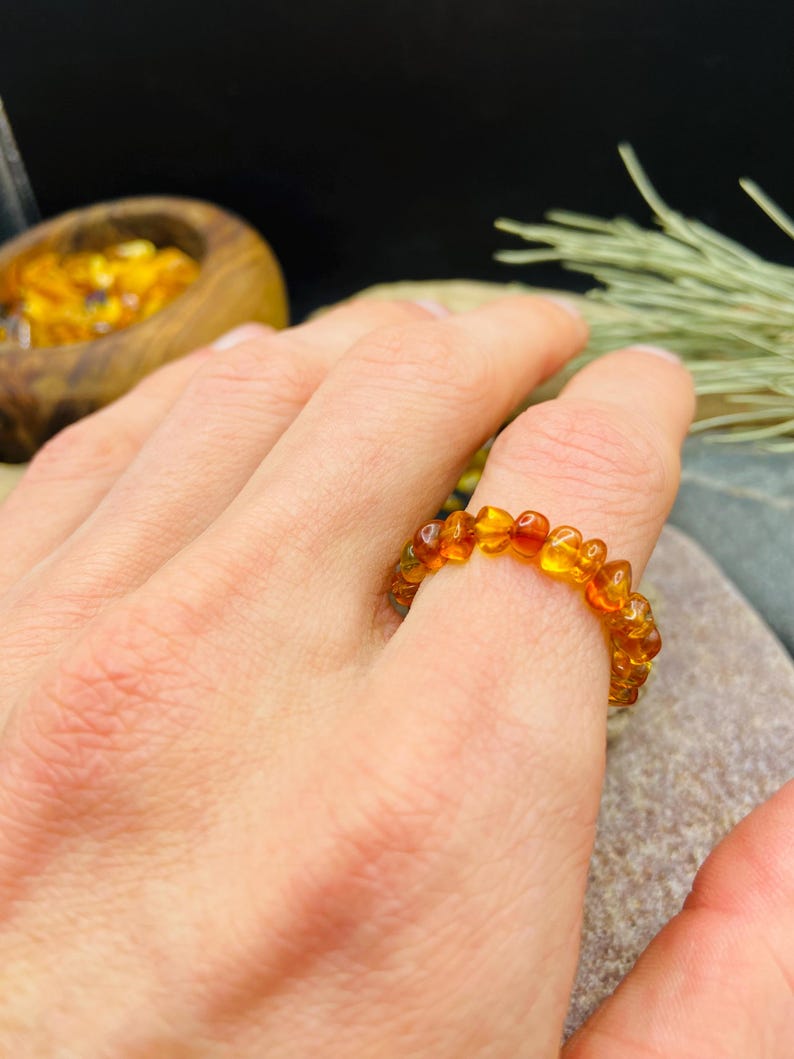 May include: A ring made of amber beads. The ring is a warm, golden-orange color and appears to be made of small, irregular-shaped beads. The ring is worn on a finger. The background is blurred, but includes a wooden bowl and greenery.