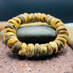 May include: A beaded bracelet composed of irregularly shaped, amber-colored beads with dark brown and cream-colored markings. The bracelet is displayed on a dark gray stone against a reddish-brown surface, showcasing its natural tones and textures.