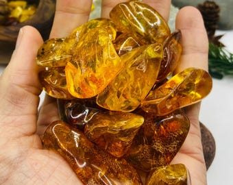 May include: A handful of polished amber stones in various shapes and sizes. The amber pieces range in color from light yellow to deep orange-brown, with visible internal patterns. The stones are held in a hand, with a blurred background.