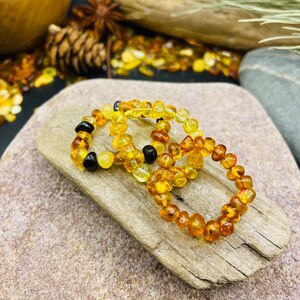 May include: Three amber beaded bracelets with varying shades of yellow, orange, and black beads. The bracelets are displayed on a piece of driftwood, with a neutral background.