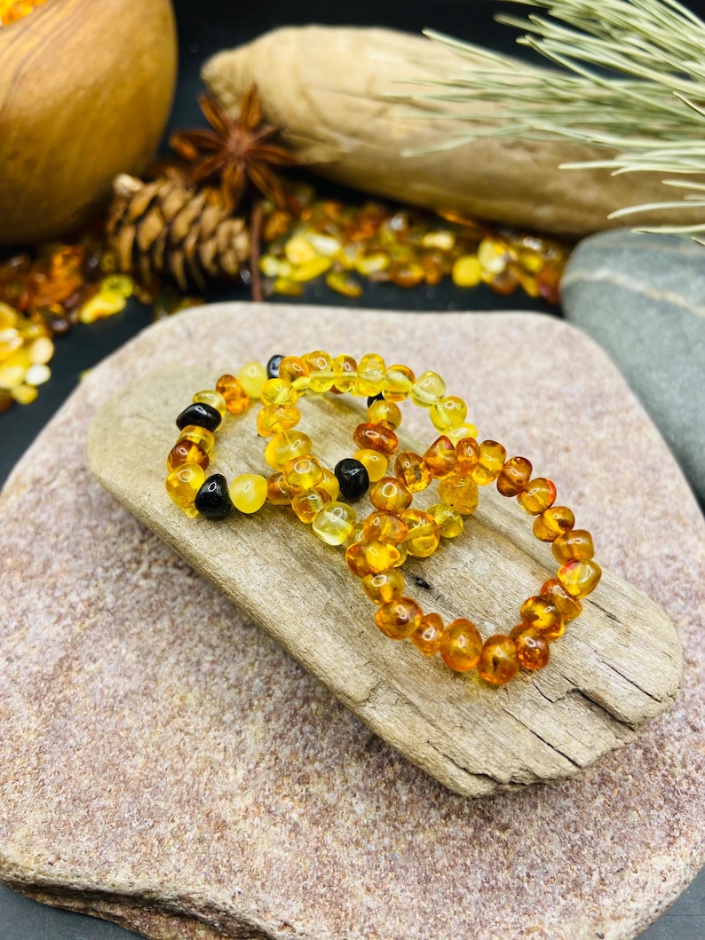 May include: Three amber beaded bracelets with varying shades of yellow, orange, and black beads. The bracelets are displayed on a piece of driftwood, with a neutral background.