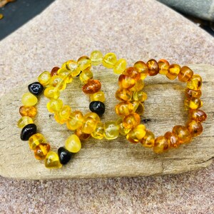 May include: Three amber beaded rings, each with a unique color variation. The rings are displayed on a light brown, textured surface. The beads range in color from light yellow to dark brown, with some black beads.
