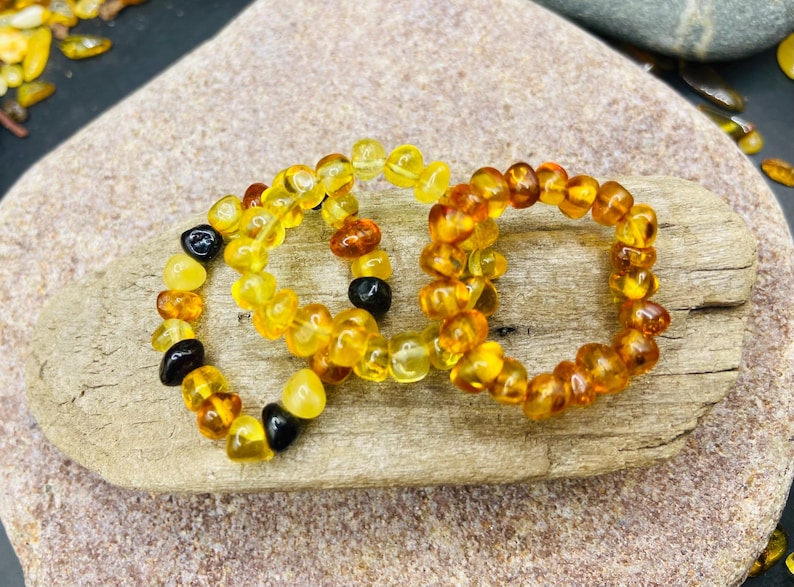 May include: Three amber beaded rings, each with a unique color variation. The rings are displayed on a light brown, textured surface. The beads range in color from light yellow to dark brown, with some black beads.