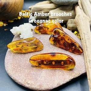 May include: Four Baltic amber brooches in cognac color are displayed on a stone surface. The brooches have a polished, translucent appearance, showcasing natural inclusions. The text "Baltic Amber brooches in cognac color" is visible.