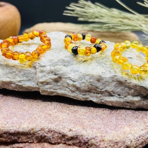 May include: Three amber rings displayed on a textured stone surface. The rings are composed of small, irregularly shaped amber beads in shades of yellow, orange, and brown. One ring includes black beads.