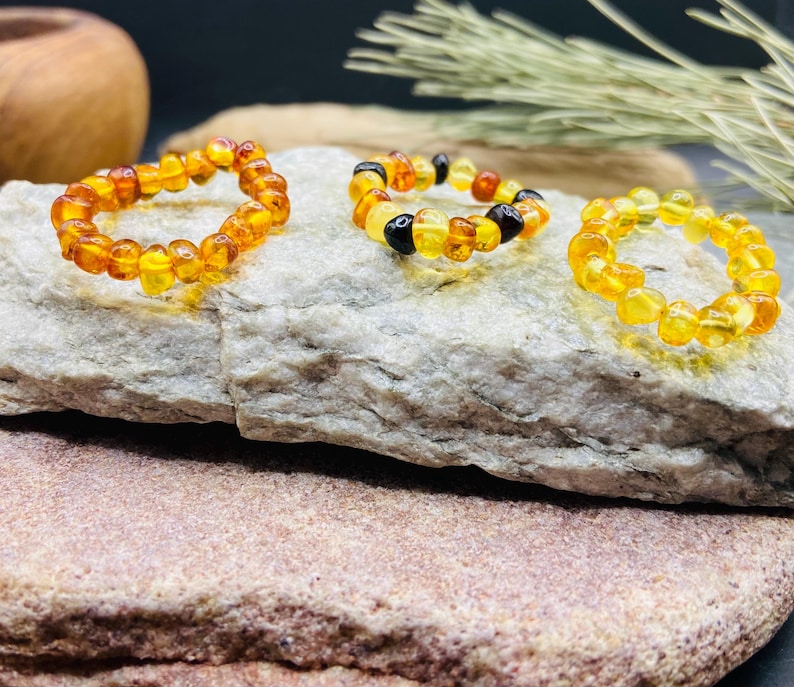 May include: Three amber rings displayed on a textured stone surface. The rings are composed of small, irregularly shaped amber beads in shades of yellow, orange, and brown. One ring includes black beads.