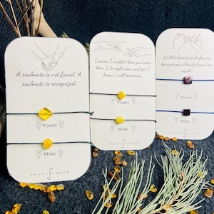 Op de afbeelding: Drie vriendschapsarmbanden met barnsteenkleurige kralen en zwarte of blauwe koorden worden gepresenteerd op kaarten met romantische citaten. De kaarten bevatten ook lijntekeningen van handen en de woorden "Yours" en "Mine."