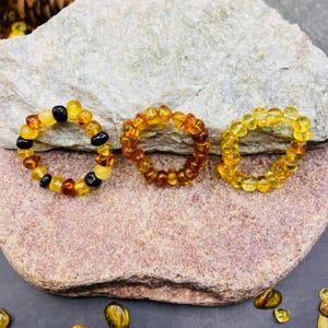 May include: Three amber rings of varying shades of yellow, orange, and brown. One ring features black beads interspersed with amber. The rings are displayed on a stone surface, showcasing the natural colors and textures of the amber.