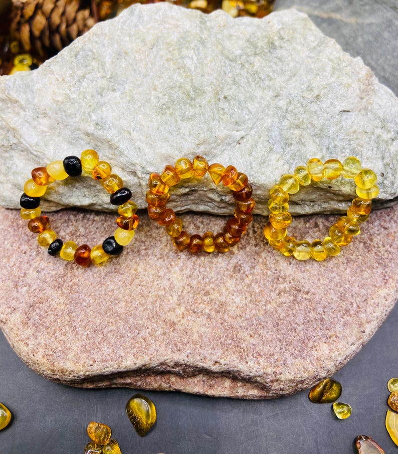 May include: Three amber rings of varying shades of yellow, orange, and brown. One ring features black beads interspersed with amber. The rings are displayed on a stone surface, showcasing the natural colors and textures of the amber.