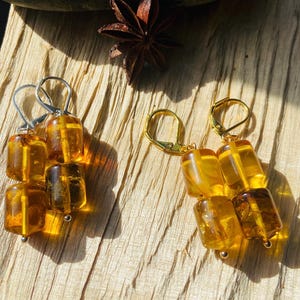 May include: Two pairs of amber earrings. One pair has silver-colored hooks, and the other has gold-colored hooks. Each earring is made of three rectangular amber stones. The earrings are displayed on a wooden surface.