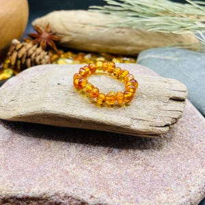 May include: A beaded amber bracelet with a warm, golden-orange hue. The bracelet is made of small, rounded amber beads strung together. The jewelry is displayed on a piece of driftwood, with a natural background.