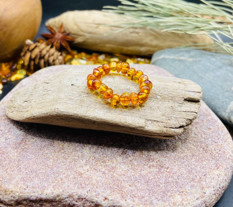 May include: A beaded amber bracelet with a warm, golden-orange hue. The bracelet is made of small, rounded amber beads strung together. The jewelry is displayed on a piece of driftwood, with a natural background.