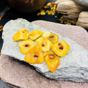 Könnte beinhalten: Eine Sammlung polierter Bernsteine in verschiedenen Formen und Größen, mit natürlichen Löchern. Die Steine haben eine warme gelbe Farbe und liegen auf einer grauen und braunen Steinoberfläche. Im Hintergrund befindet sich eine Holzschale mit Bernstein.