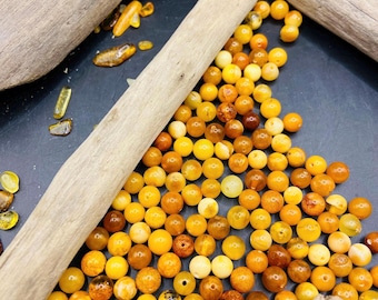 May include: A collection of amber beads in various shades of yellow and brown, scattered on a dark surface. Several pieces of driftwood are arranged around the beads. The beads vary in size and color, creating a natural, earthy aesthetic.
